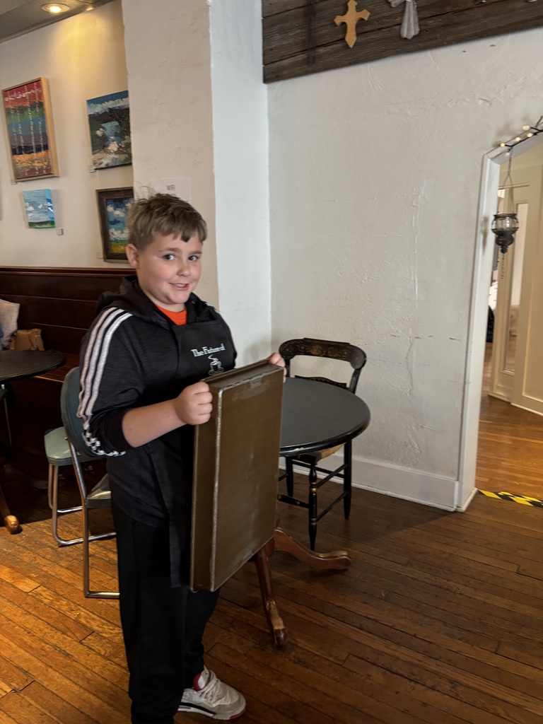 A young boy in a black jacket holds a black menu board in a room with wooden floors and white walls.