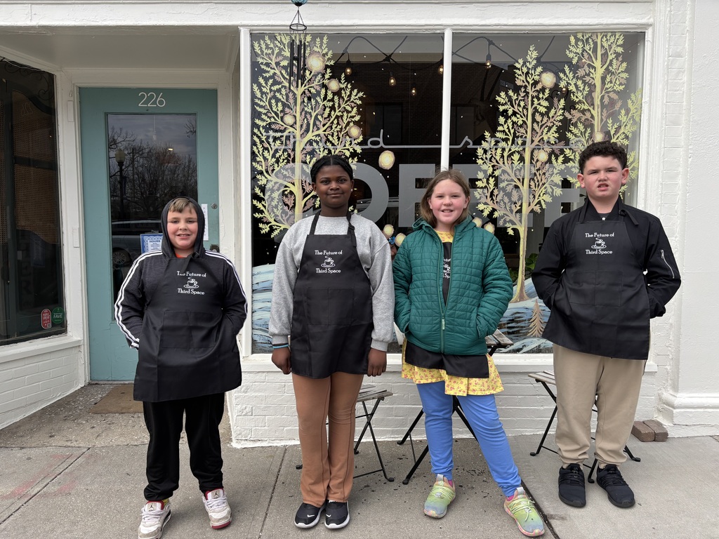Four individuals wearing jackets stand in front of a building with glass windows and a tree decoration.