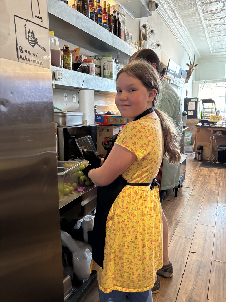 A young girl in a yellow dress and apron stands at a counter inside a kitchen.