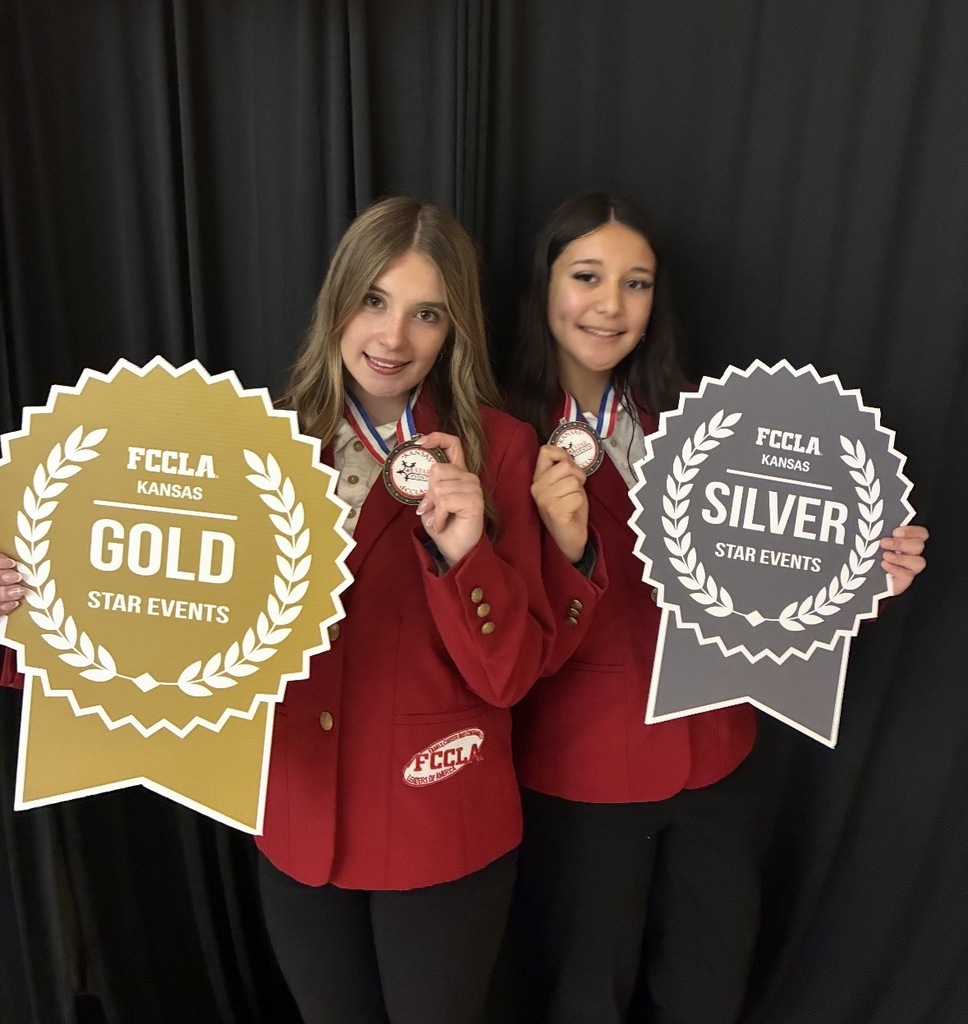 Two women wearing red jackets and black pants hold medals and signs, one reading "Gold" and the other "Silver."