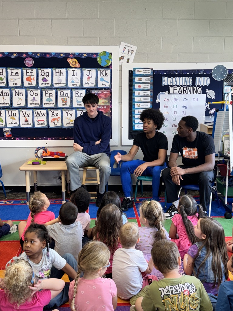 Three adults seated in front of young students in a classroom, with a colorful alphabet chart on the wall.