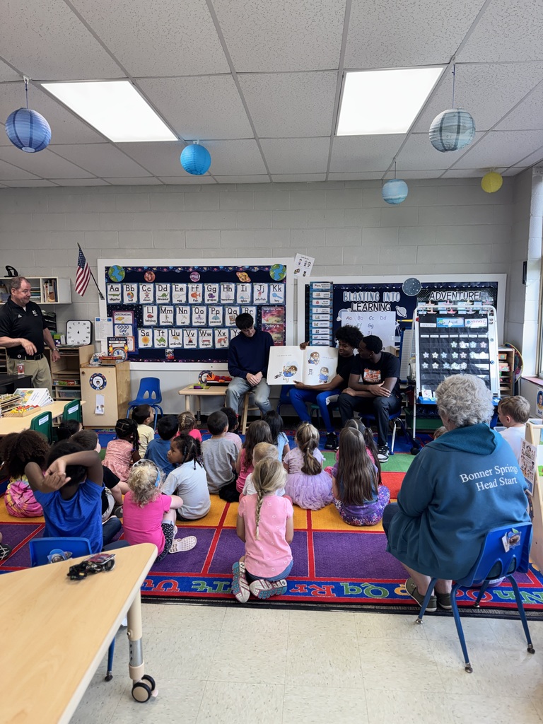 Kids sit on a floor rug while basketball players read aloud. 