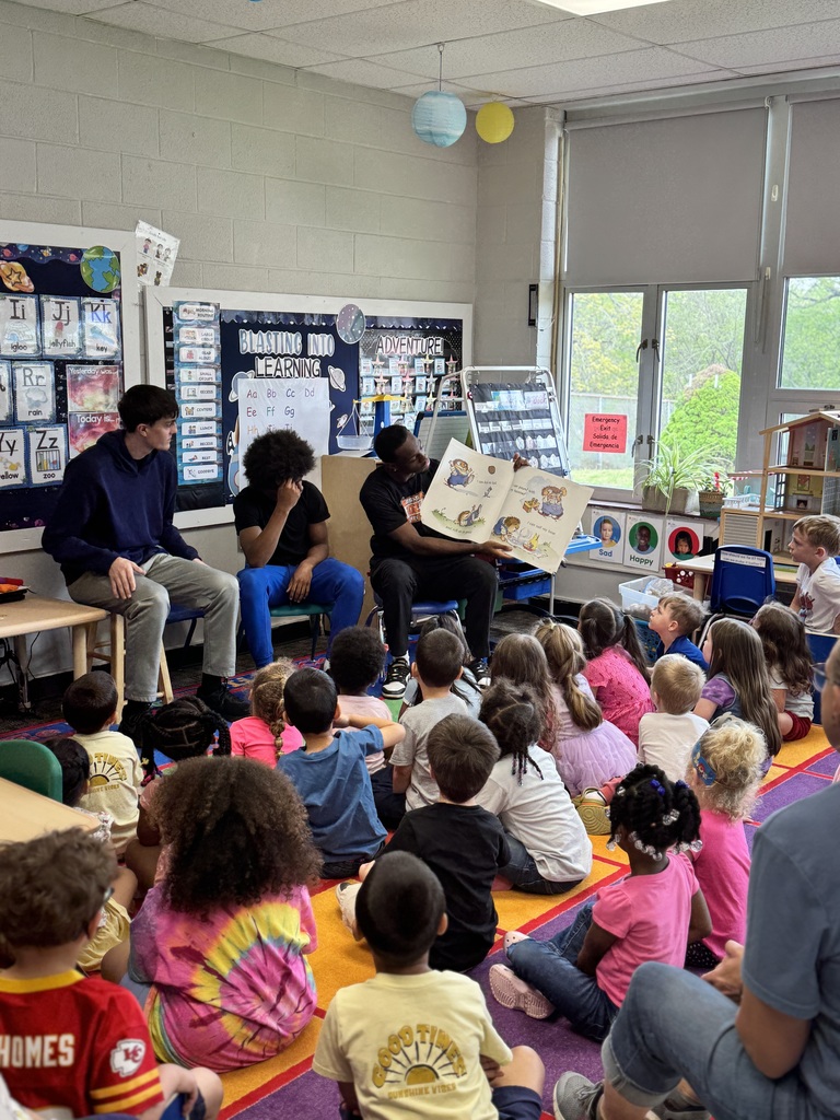 Several adults and children in a classroom, with adults reading a book to the children.