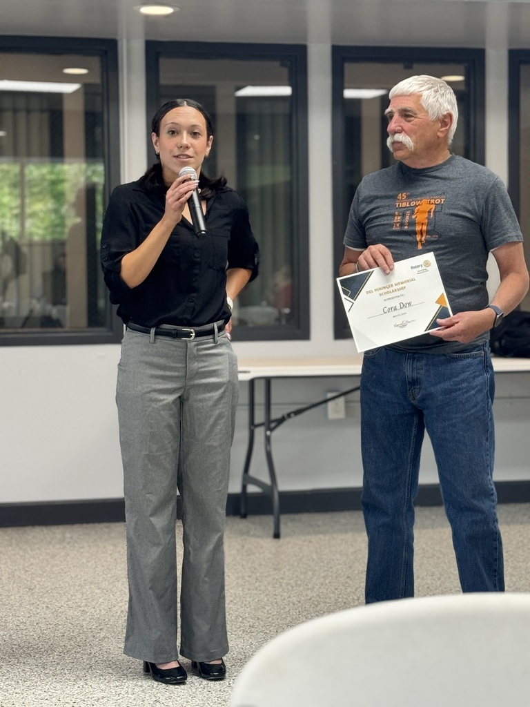 A woman with a microphone stands with a man holding a certificate, in front of a table and windows.