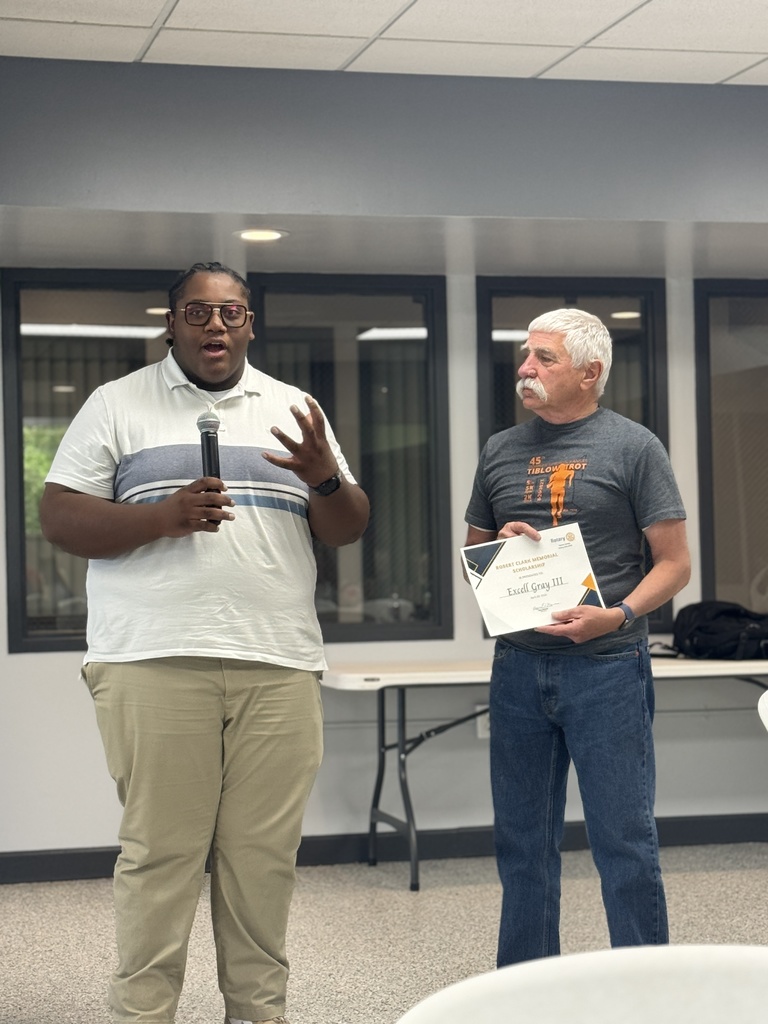 Two men in a room with windows; one holds a microphone and speaks while the other holds a certificate.