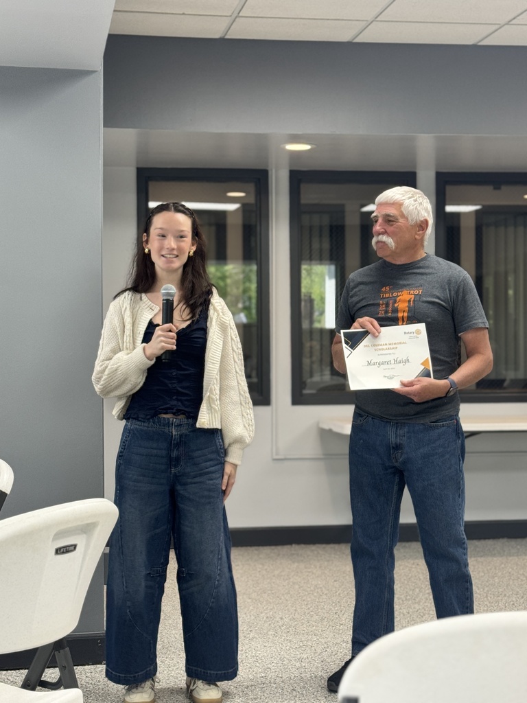 A woman with a microphone stands next to a man holding a certificate, both wearing casual clothes, in a room with chairs and windows.