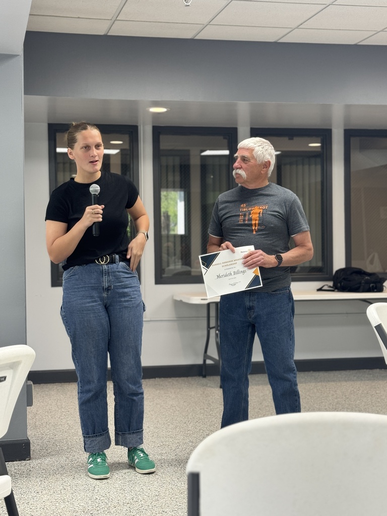 A woman holds a microphone and a man holds a certificate in a room with chairs and a table.