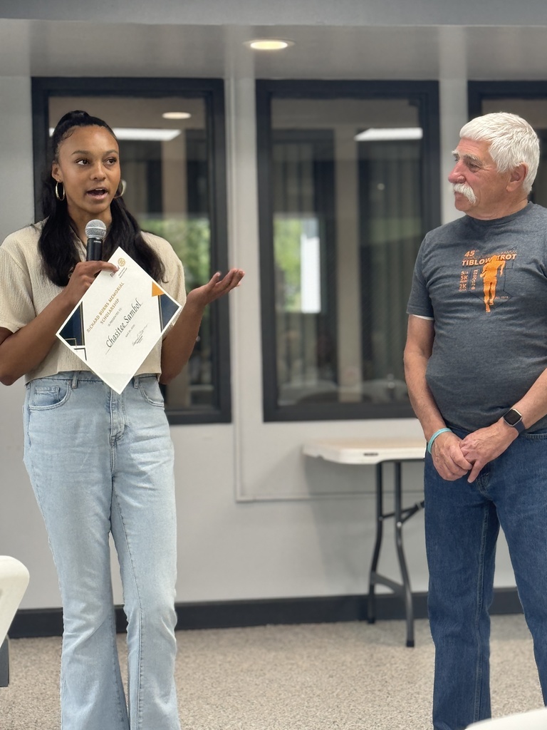 Woman holding a certificate and microphone stands in front of a man in an indoor room.