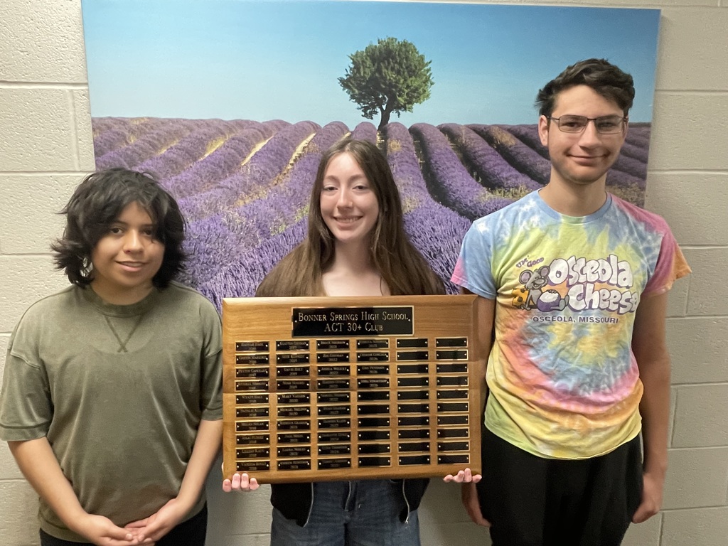 Three individuals pose next to a large plaque with text. A woman holds it between two people. A field of lavender flowers is visible in the background.