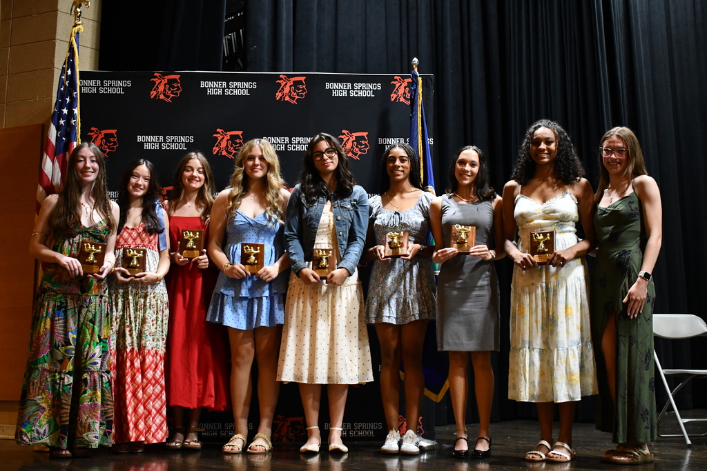 A group of women holding awards, lined up on a stage, with a flag and black backdrop.