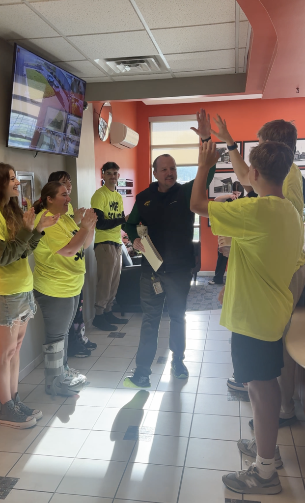 People in yellow shirts and sneakers stand in a hallway. A man in black is giving high fives.