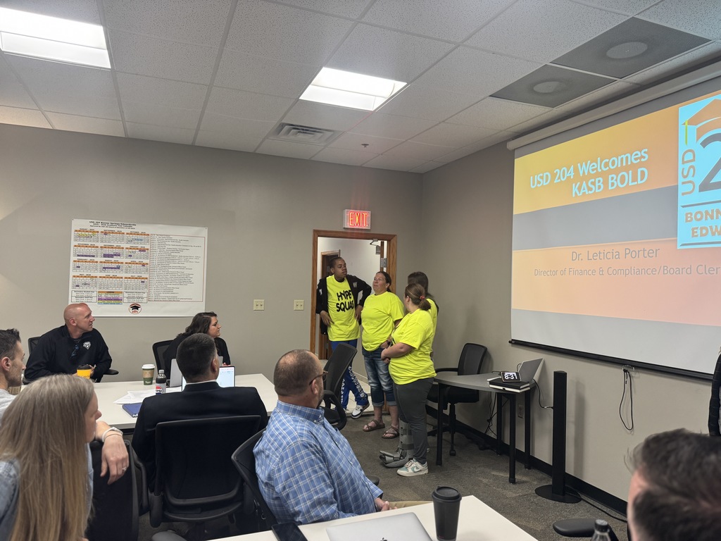 A room with a projector screen displaying "2020 welcomes KASB BOLD". People are seated at tables.