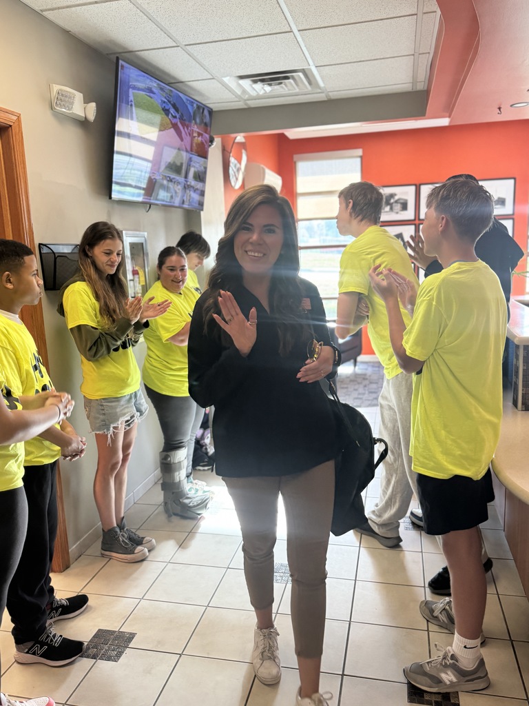 A group of people in yellow shirts are standing in a tiled room. A woman in black stands in the middle, clapping.