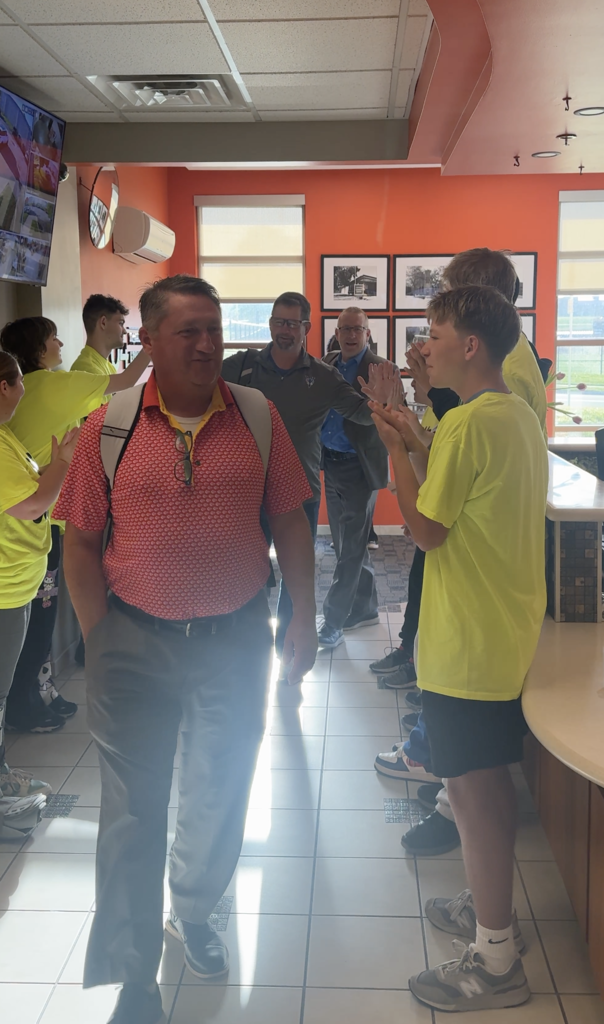 A group of adults and children wearing yellow shirts stand in a hallway with orange walls and white tile floor.