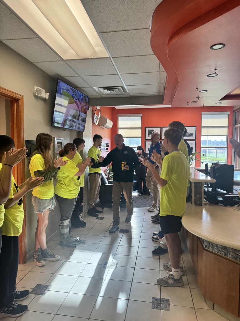 A group of people in yellow shirts stand in a lobby, clapping and facing a man speaking. Bright lighting, tiled floor, and a television are visible.