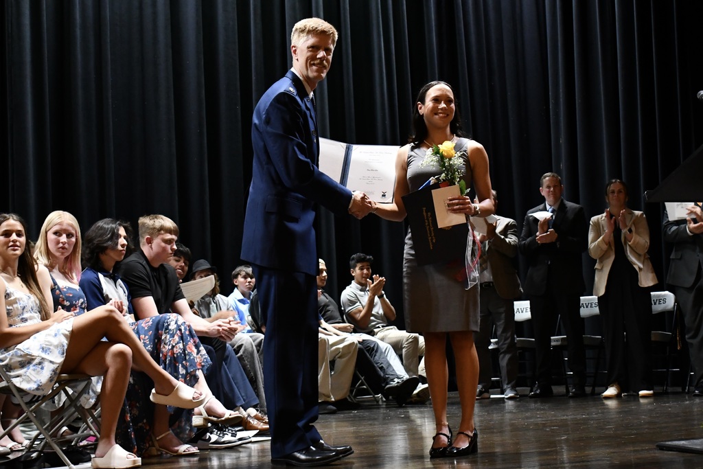 A man and a woman stand on stage, holding a paper and flowers. Behind them, people sit and stand, observing.