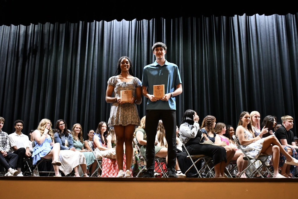 Two individuals stand on a stage holding awards. Behind them, an audience sits in rows of chairs, watching.