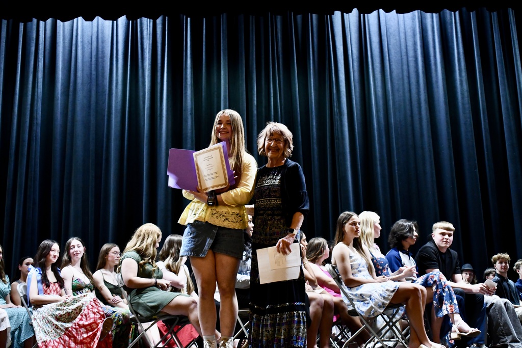 Two women stand on stage at an event, one holding a purple folder, the other holding papers. Behind them, seated audience members watch.