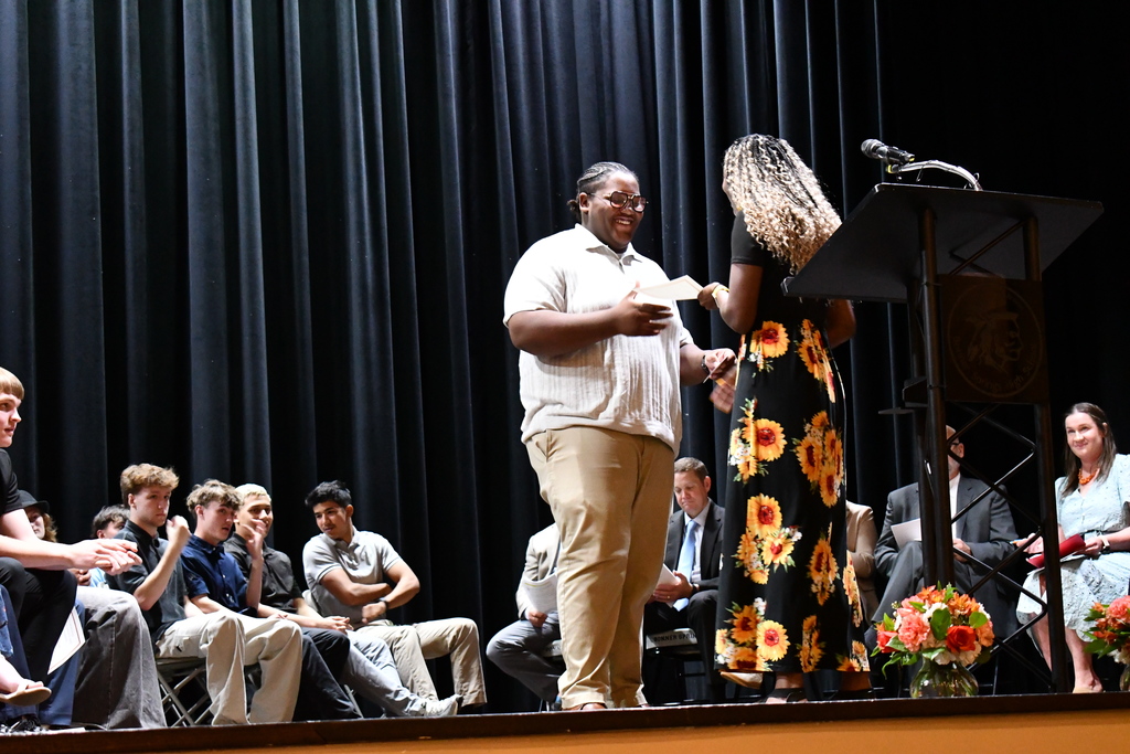 A person on stage holding a paper and another person handing it to them. Behind them is a podium with a microphone and flowers.