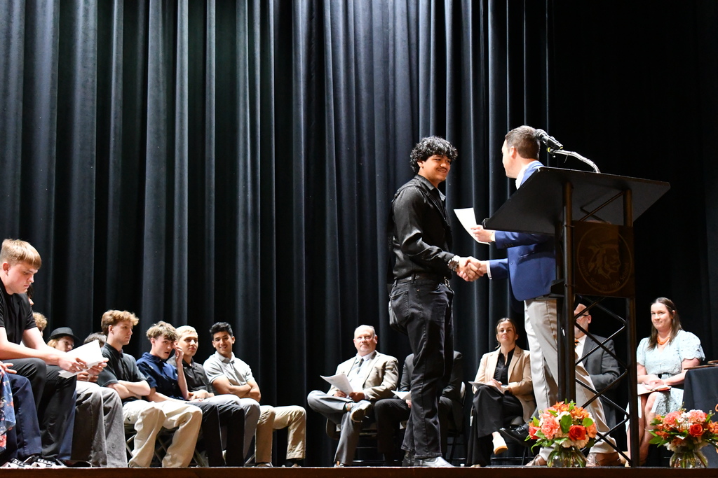 On a stage, two individuals shake hands. One holds a paper, and a microphone is nearby. Behind them, people sit on chairs, and a black curtain is in the background.