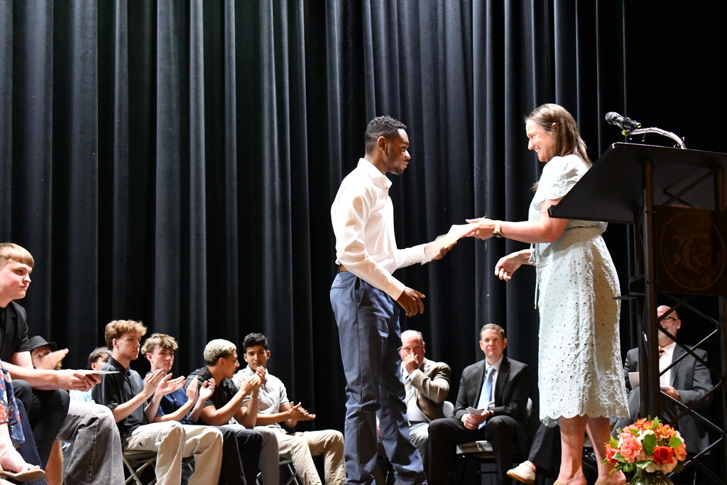 People on a stage, one in a dress, one in a suit, shaking hands. Behind them, a microphone on a stand and flowers.
