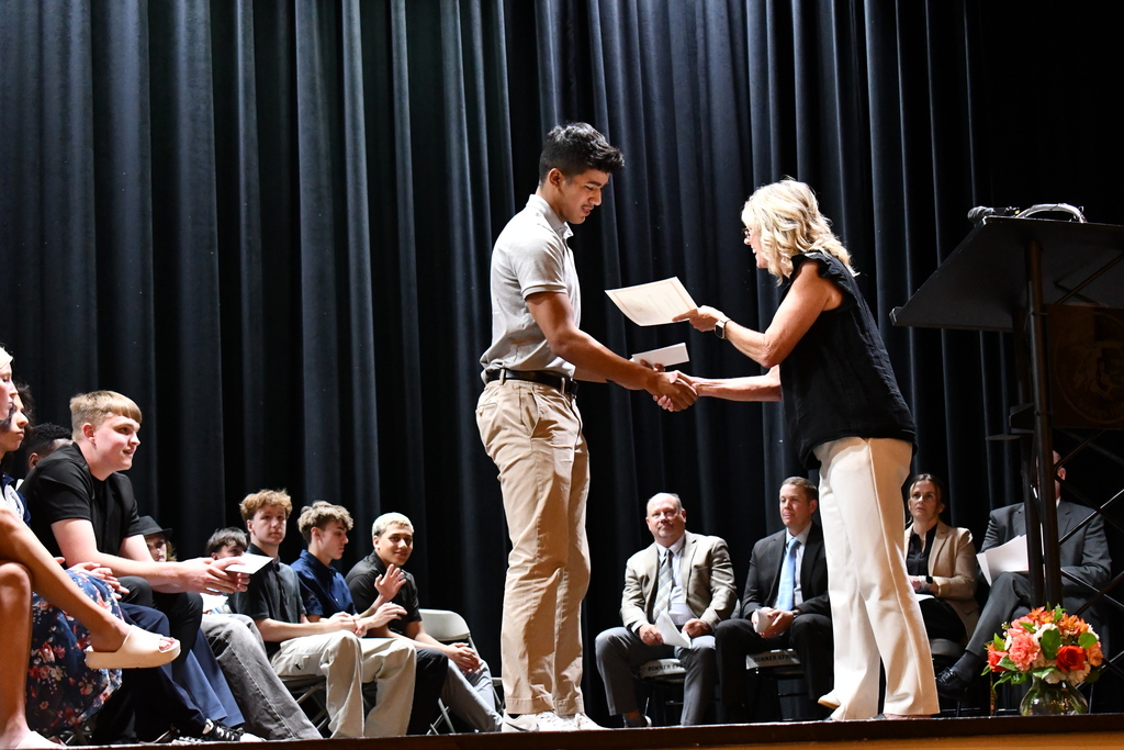 A man and a woman on stage shake hands while holding papers; people sit in the audience.