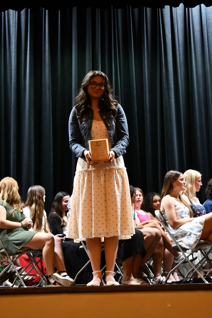 A person stands on a stage holding an award while others sit in chairs, some wearing glasses.