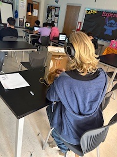 A classroom with students seated at desks, a woman wearing headphones and using a laptop, and a chalkboard with colorful drawings.