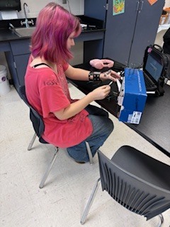 Woman in red shirt and jeans seated, working on a blue box, with a laptop nearby.