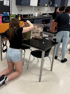 Two people are in a classroom, one leaning over a table with a box on it.