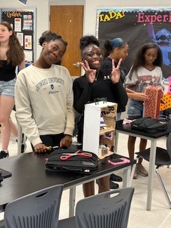Three students pose in a classroom. One makes a peace sign with both hands. The table in front has a backpack and a small box.