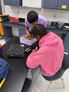Two students in a classroom setting, one in a pink hoodie, the other in a purple shirt, both working on a box.