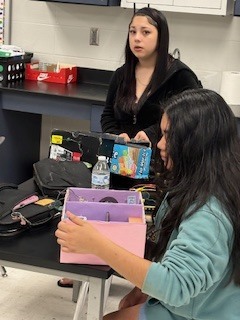 Two people in a classroom setting, one holding a box, the other standing, surrounded by various items.