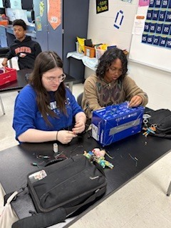 Two students seated at a desk, one wearing glasses, work on a project with various materials.