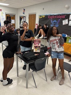 Three women stand in a classroom, with one making a peace sign. They are in front of a desk with a laptop, while others sit on chairs behind them.