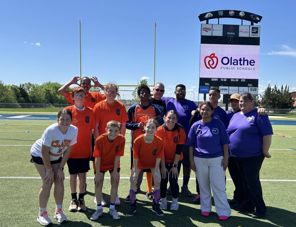A group of people in orange and white shirts stands on a field, with a scoreboard and sign behind them.