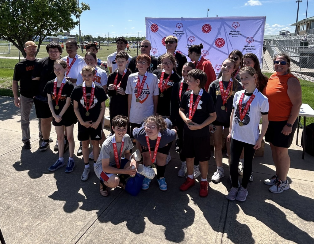 A group of children, adults, and two banners stand on a concrete path with grass and trees.