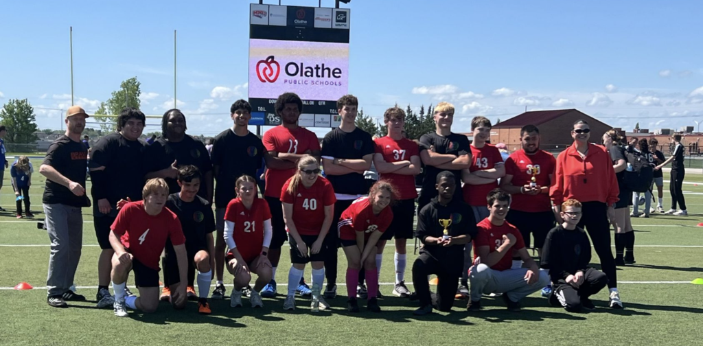 A team in red jerseys stands together on a field with several people behind them, under a bright sky.