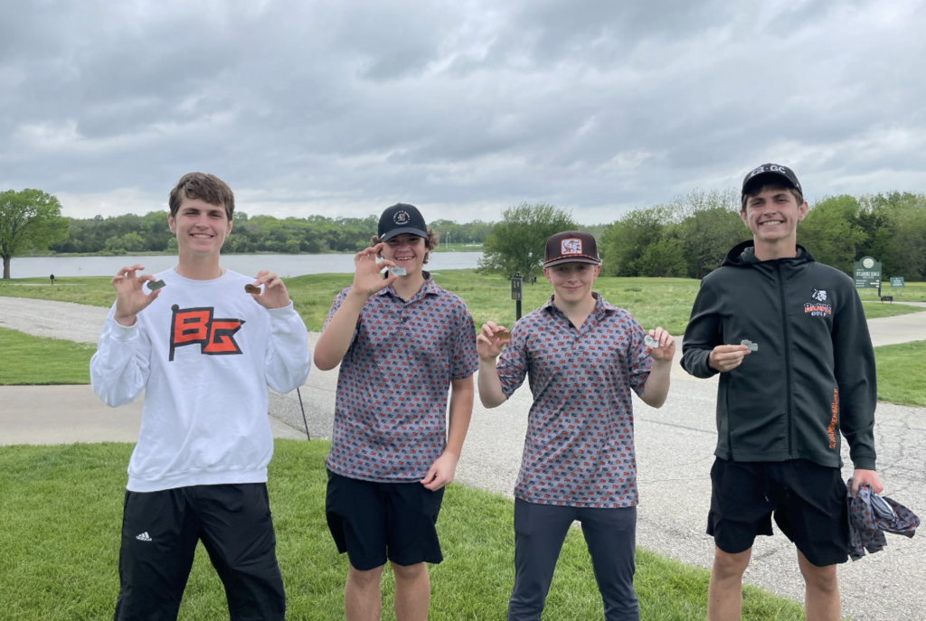 Four individuals stand on grass, holding medals. Two wear hats, and two wear jackets. A water body is behind them.