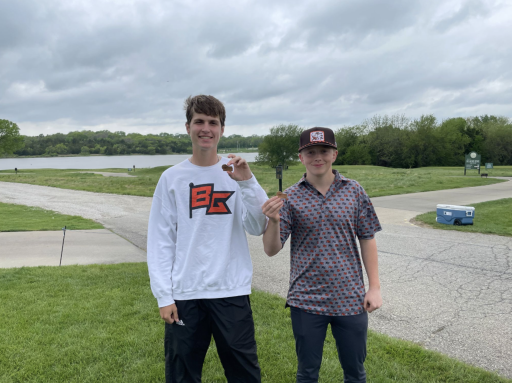Two young men stand on a golf course, one in a white shirt and the other in a cap.