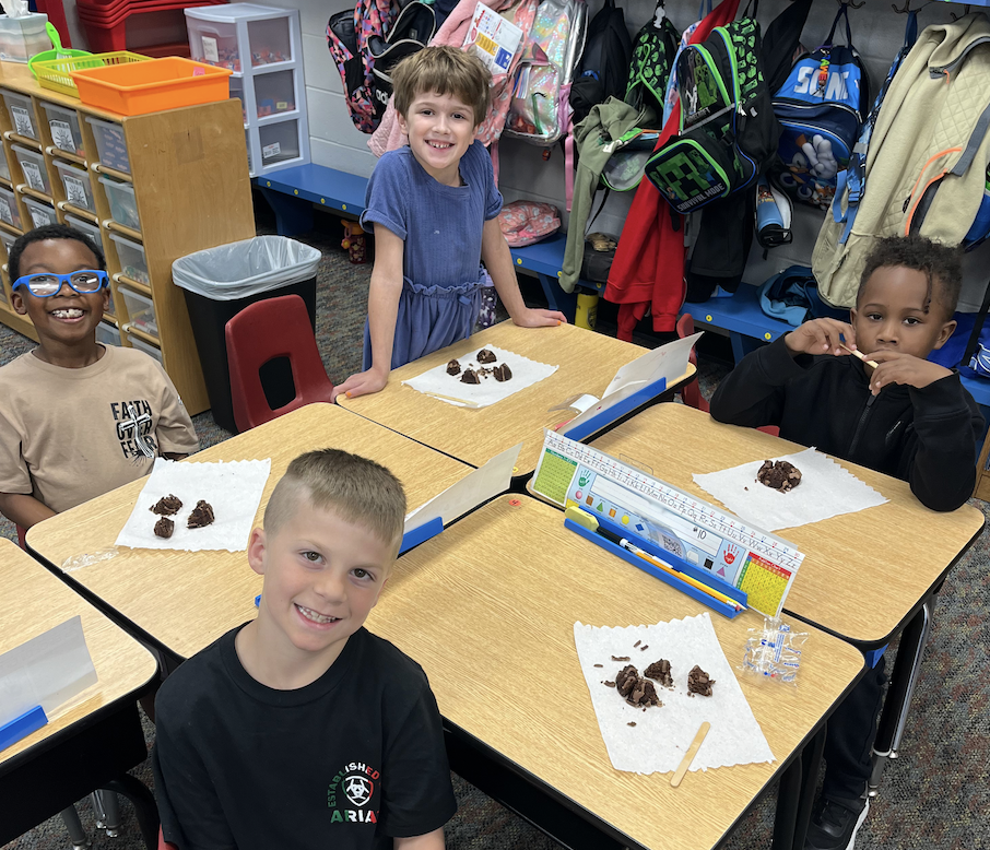 Four children sit at a desk with chocolate pieces on paper. They are smiling. Behind them, a classroom is visible with backpacks and storage boxes.