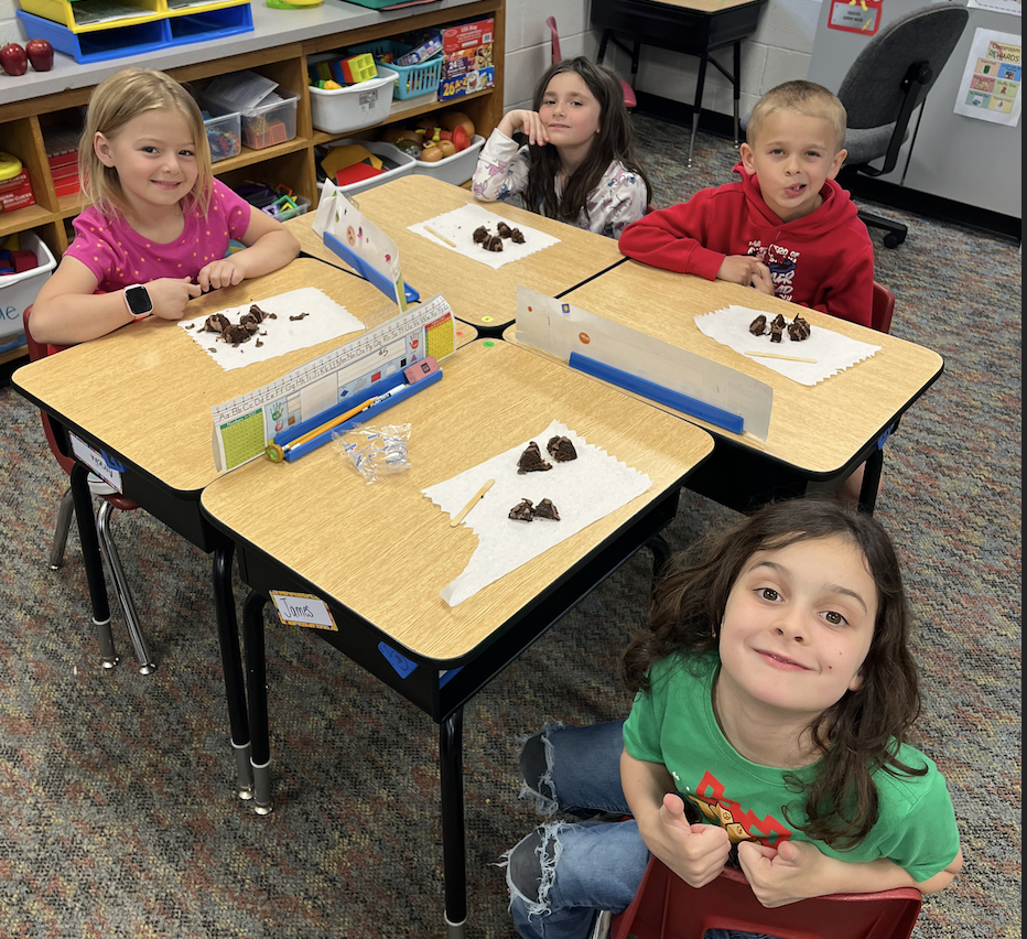 Four children sit at desks, each with chocolate chips on a paper. They smile at the camera.