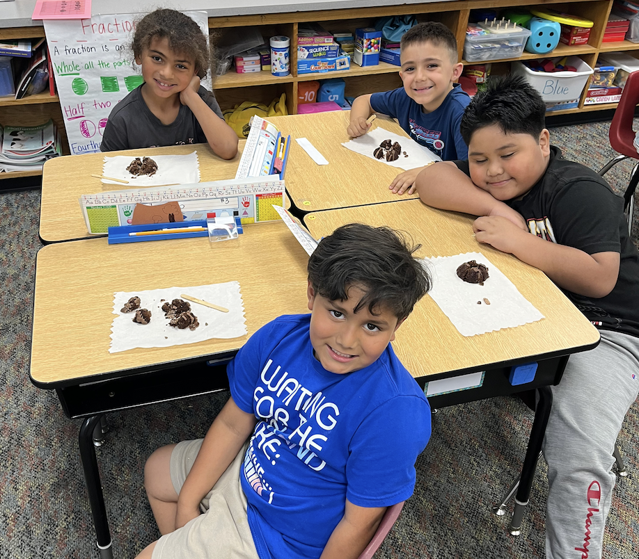 Four children sit at desks in a classroom, engaged in an activity. Three are smiling, one appears thoughtful.