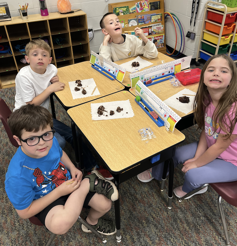 Four children seated at desks, two boys and two girls, with food on their desks. They are smiling.