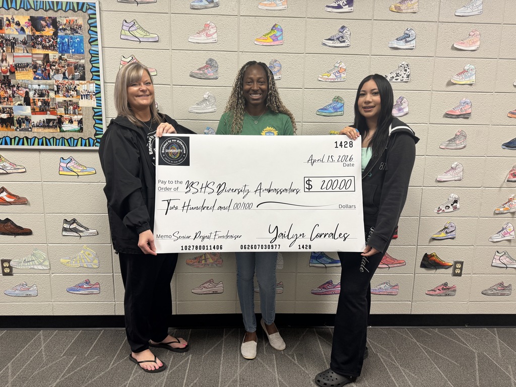 Three women hold a check in front of a wall covered with pairs of sneakers.