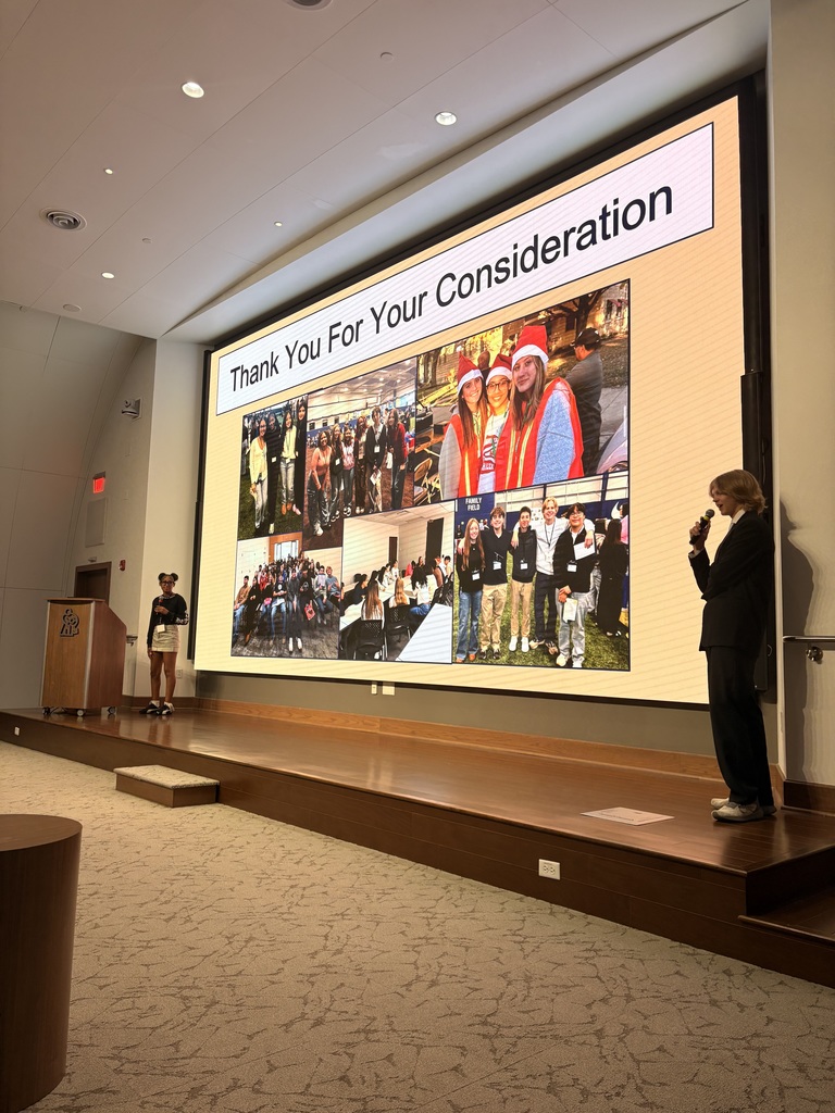 A person stands on stage with a microphone while a collage of photos plays on the screen.