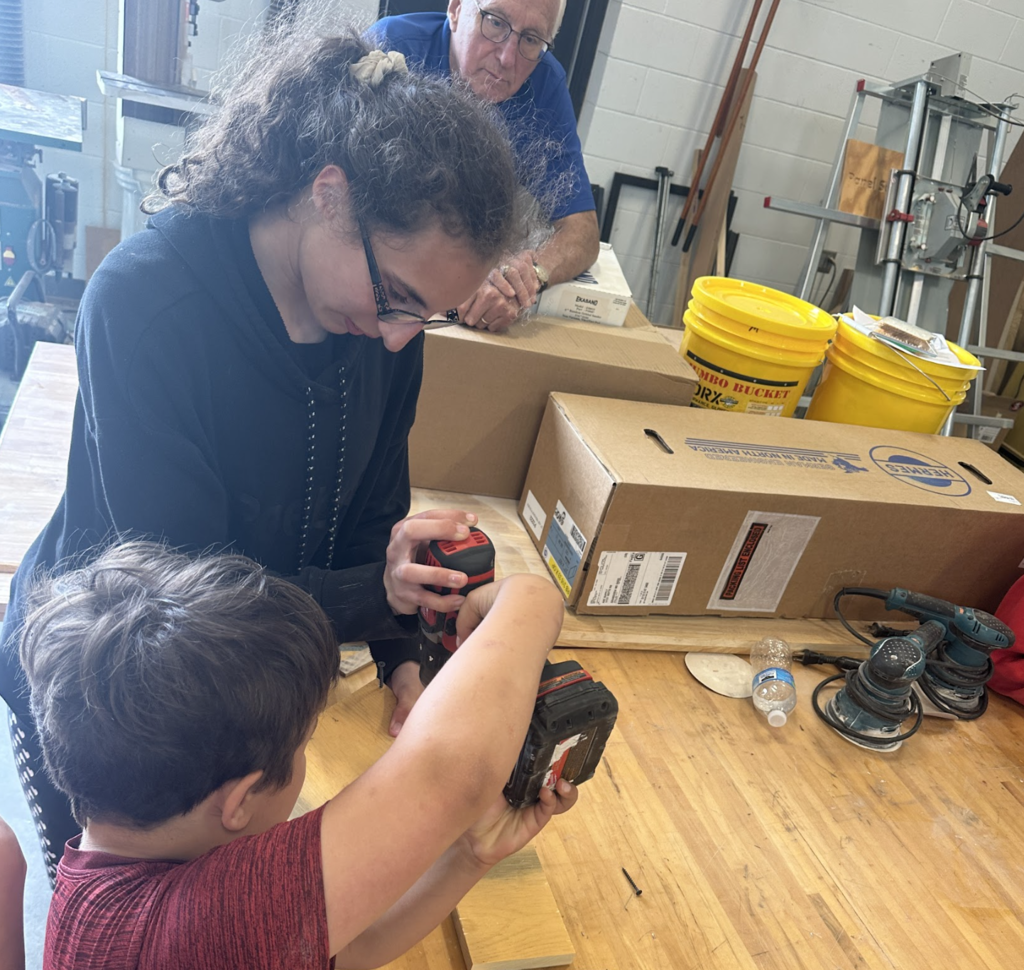 A woman helps a child with a drill, surrounded by wooden planks and tools in a workshop.