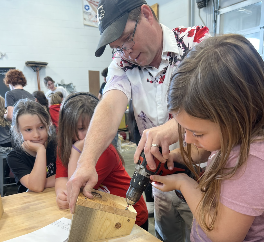 A man helps two girls drill into a wooden box. One girl wears a pink shirt, another black, and the third red.