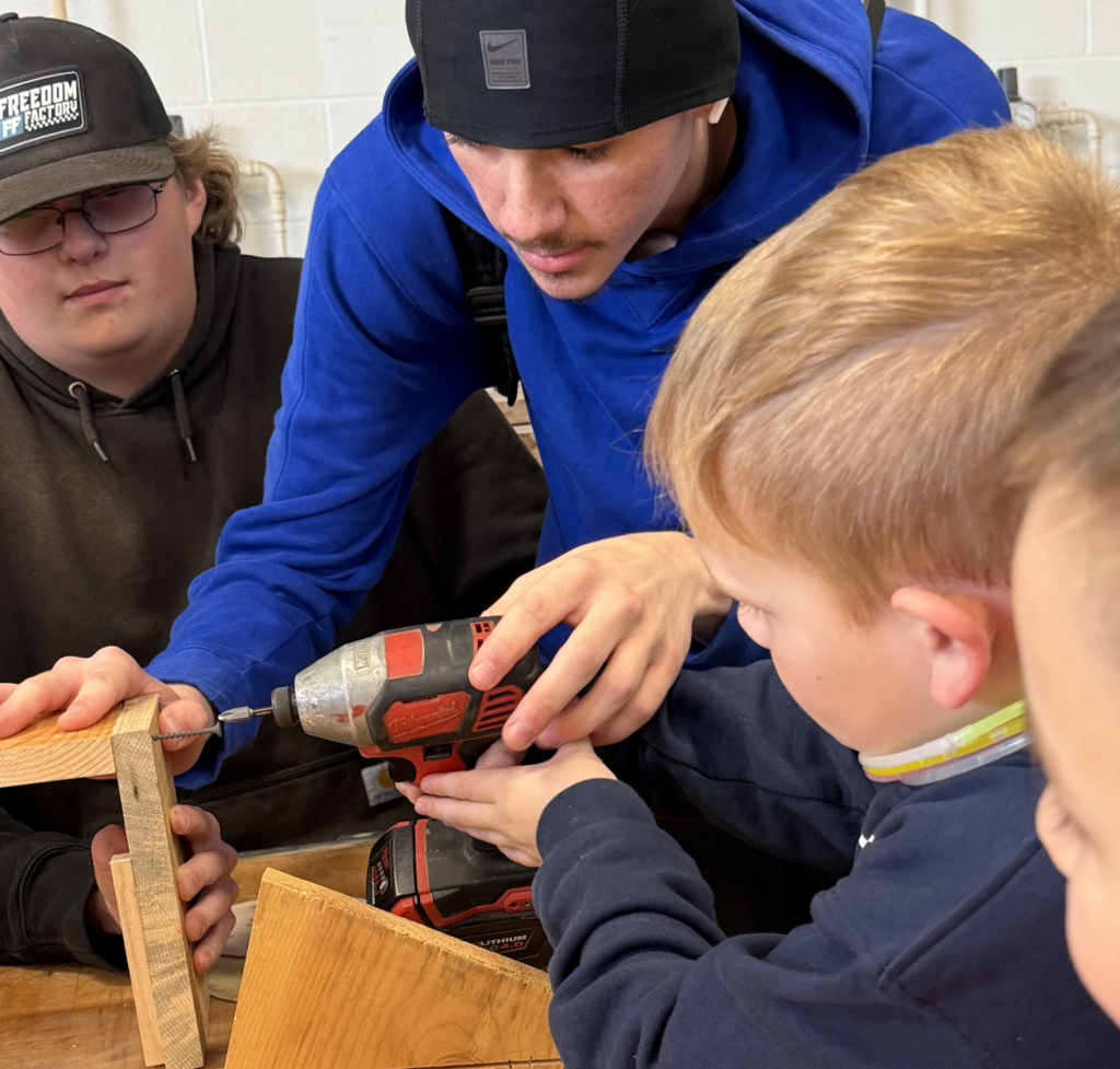 Three people in a workshop. A man uses a drill, another man guides his hand, and a boy watches.