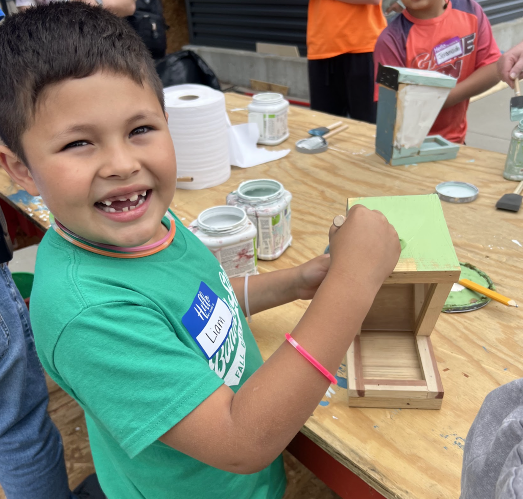 A child with a green shirt smiles while painting a wooden box. Behind, others work on similar boxes.